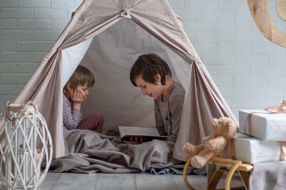 Two siblings reading books in a reading nook set up inside a tent.