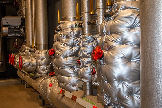 Industrial pipes and valves with silver insulation jackets in a mechanical room.