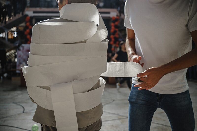 Child wrapping another in toilet paper during a mummy race at a trunk or treat event. The person being wrapped is nearly covered, with Halloween decorations and other participants in the background.