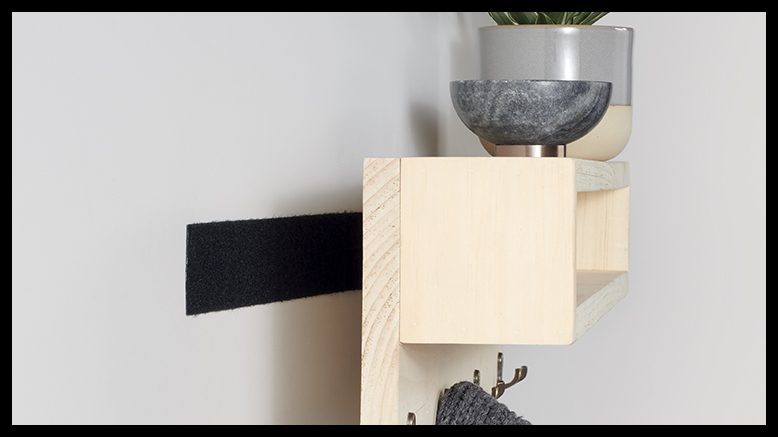 Wall-mounted shelf secured with VELCRO® Brand Industrial Strength Tape for home improvement, featuring a potted plant on top and hooks below holding towels or cloths.