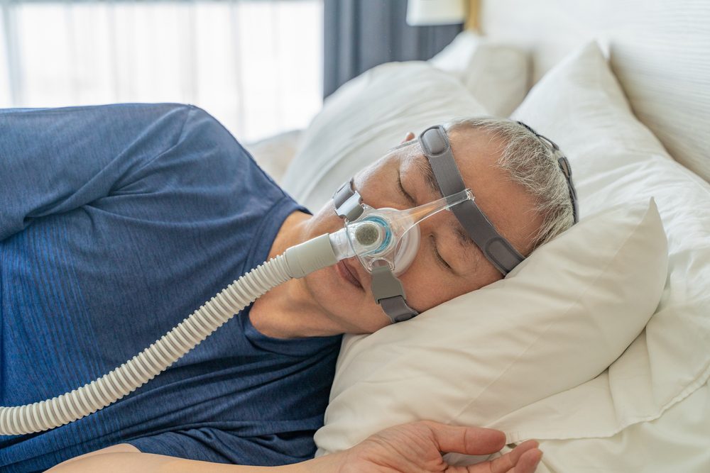 Person with short gray hair lying on a bed, wearing a blue shirt and using a CPAP machine with VELCRO® Brand DFL fastener.