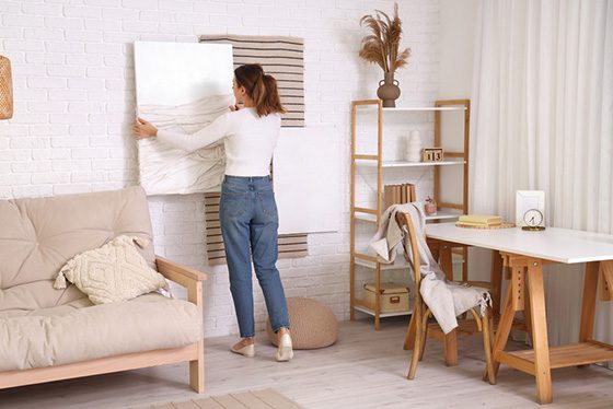 Woman adjusting a large canvas painting on a white brick wall using VELCRO® Brand fasteners in a cozy living room with modern decor, including a beige sofa, wooden shelving unit and desk.