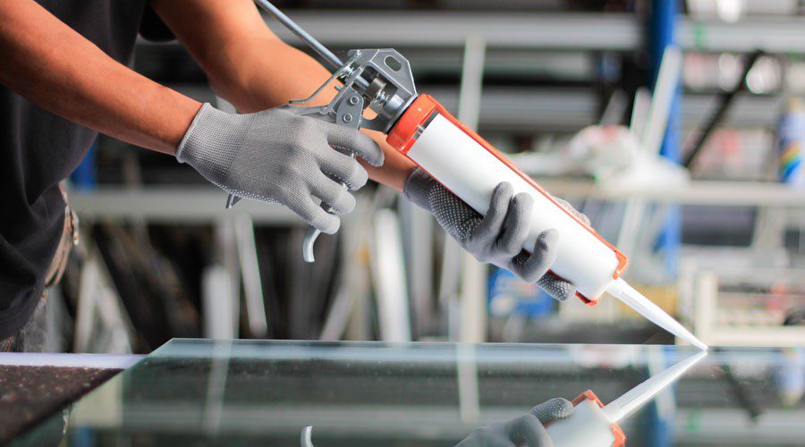 Worker applying adhesive with a caulking gun in a manufacturing setting, highlighting the mess and chemical exposure risks compared to clean, high quality hook and loop fasteners with pressure sensitive adhesive options