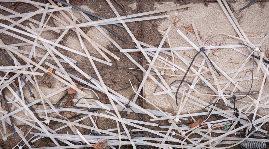 Discarded white and black plastic zip ties scattered on the ground, illustrating the waste created by single-use fasteners and the need for reusable hook and loop alternatives in the manufacturing process.