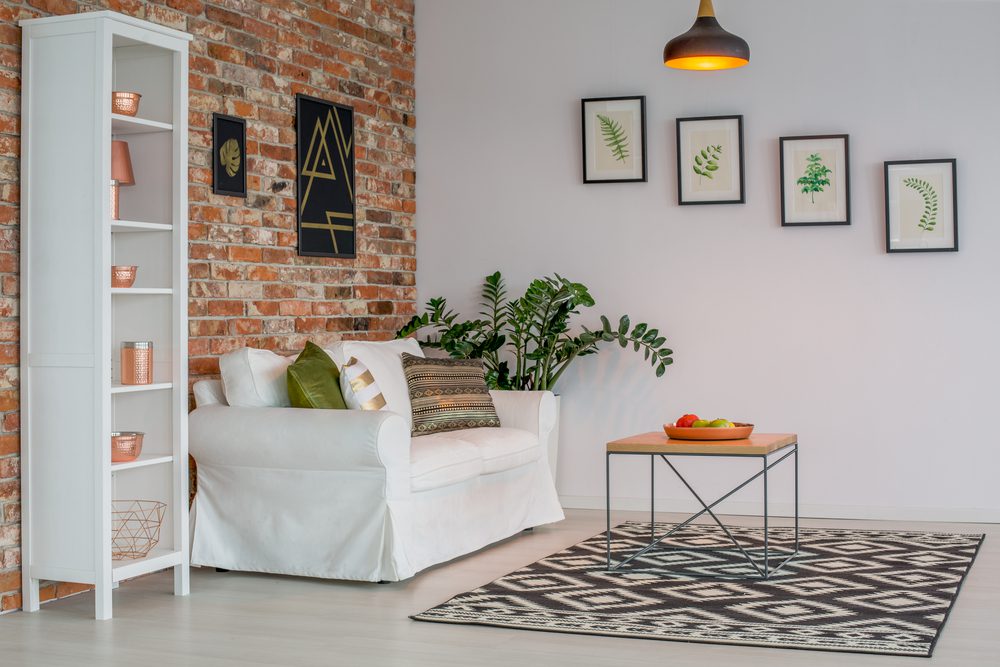 Stylish modern living room with a white sofa and green cushions set against a brick wall, decorated with geometric artwork and framed botanical prints. A wooden coffee table sits on a black and white rug, with VELCRO® Brand fasteners used to mount wall décor without drilling.